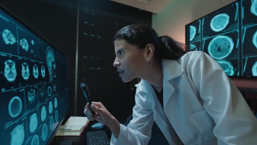 A female neurologist in a white lab coat carefully analyzes brain scans on computer monitors in a dark, high-tech medical office