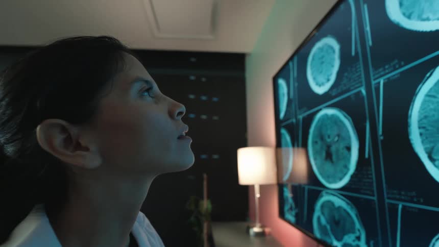 A dedicated female doctor in a dark room analyzes complex brain scans on a large digital display