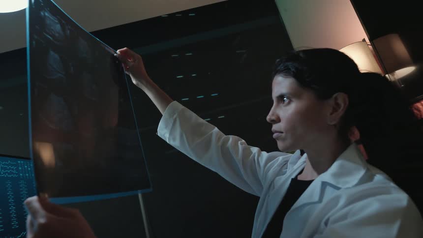 A female doctor in a lab coat intently examines a brain scan X-ray, comparing it to digital images on her computer screen in a dimly lit laboratory
