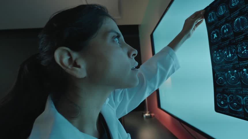 A female doctor in a dark room holds up a brain scan to a large light screen for analysis