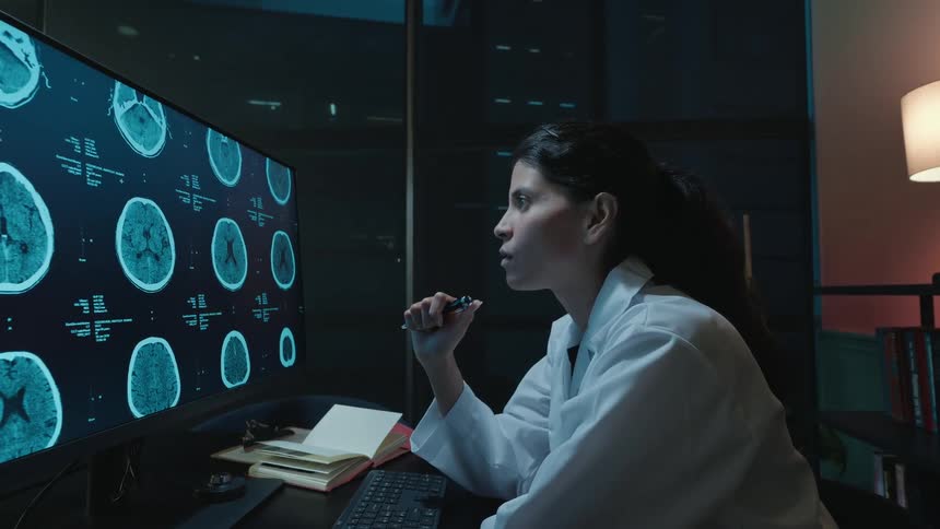 A dedicated female medical researcher analyzes a patient's brain scans on a large monitor in her city office late at night