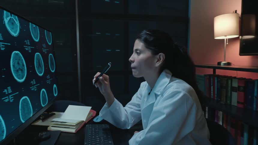 A dedicated female scientist works late into the night, studying detailed brain scans on a computer monitor