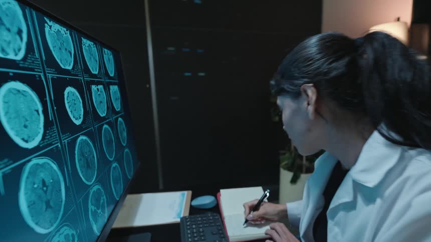 A medical professional carefully studies a series of brain scans on a large monitor before documenting her findings in a notebook