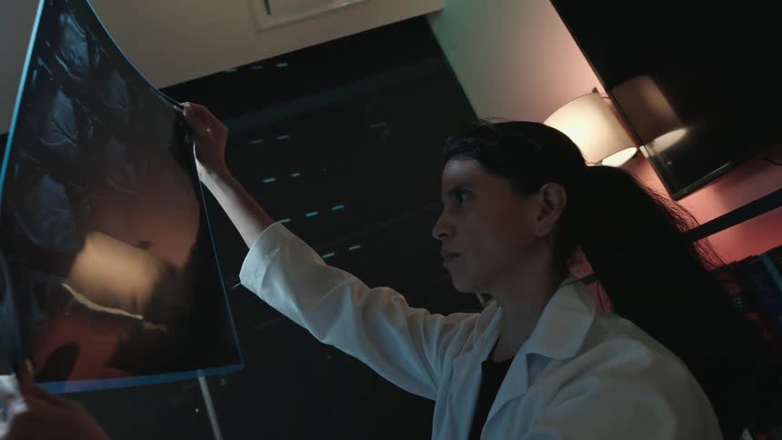 Slow motion side view of a female doctor in a white lab coat intently examining an x-ray in a dark room