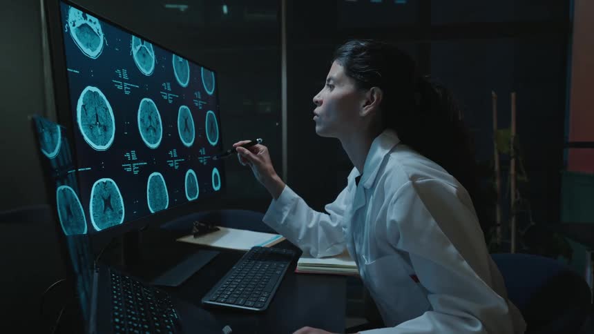 A dedicated female doctor in a lab coat meticulously examines brain scans on a computer monitor while working late in her office