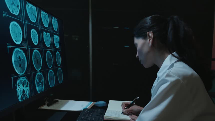 A female radiologist in a dark room meticulously studies a patient's CT brain scans on a monitor and writes down her findings