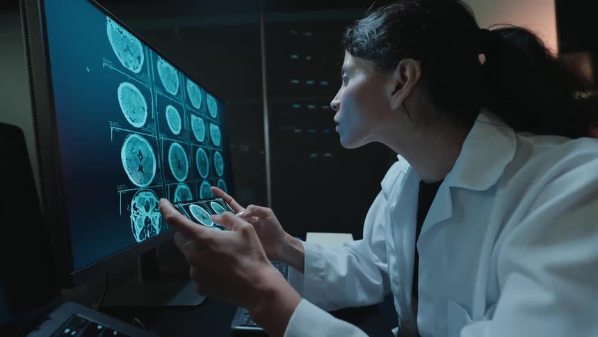 A dedicated female doctor meticulously examines brain CT scans on her computer and phone, working late into the night in a modern medical office
