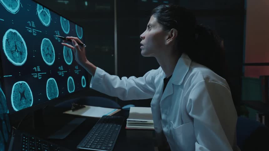 A focused female doctor in a lab coat analyzes CT brain scans on a large monitor while working late in her office