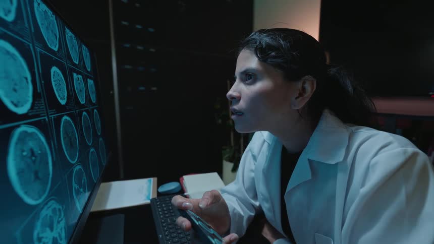 A female doctor intently analyzes brain scans on a computer monitor in a dimly lit office, using her smartphone for reference and showing a moment of realization