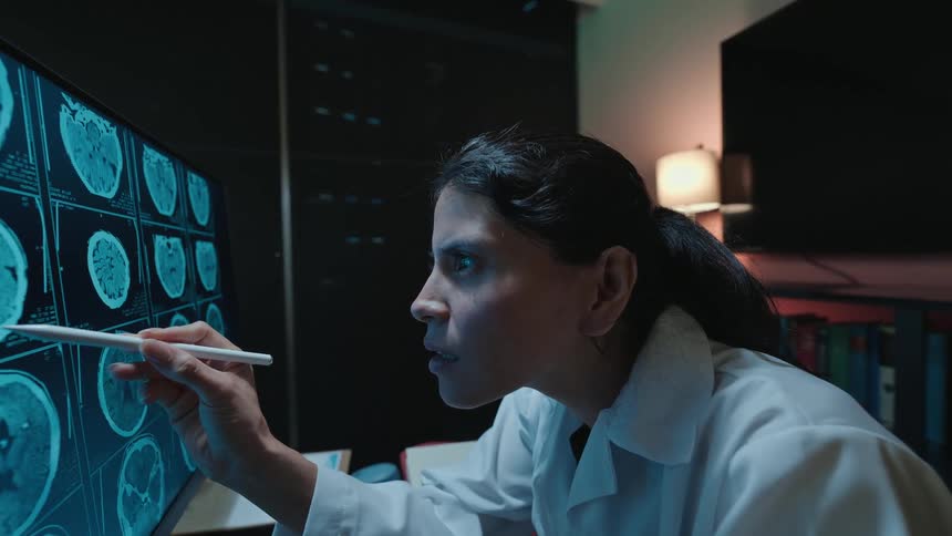 A female medical professional carefully analyzes a patient's MRI brain scans on a computer screen in a dark room