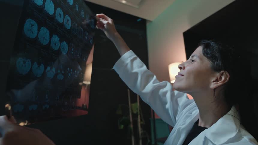 A dedicated female doctor examines a patient's brain MRI scan in her dimly lit office, comparing it with digital images on a monitor