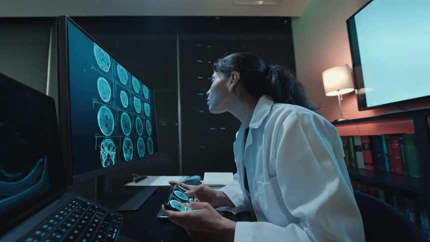 A focused female doctor in a lab coat analyzes CT brain scans, comparing images on her smartphone and a large computer monitor in a modern, dimly lit office