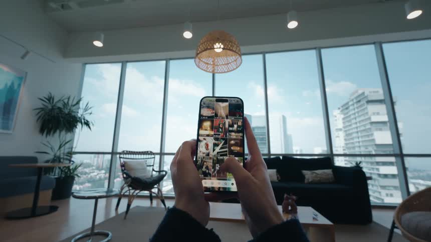 A first-person view of hands holding a smartphone and scrolling through a social media feed in a modern apartment overlooking a cityscape