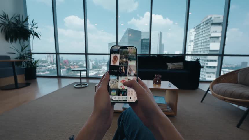 POV of a person scrolling through a social media photo feed on a smartphone while relaxing in a modern city apartment with panoramic windows