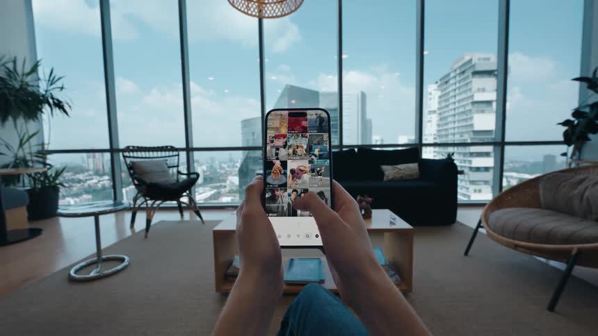 First-person perspective of a person scrolling through a social media feed on a smartphone in their modern apartment with a city view