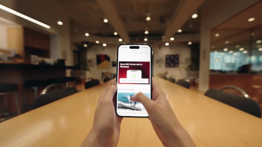 A person's hands hold a smartphone and scroll through an online news feed while sitting at a long table in a conference room