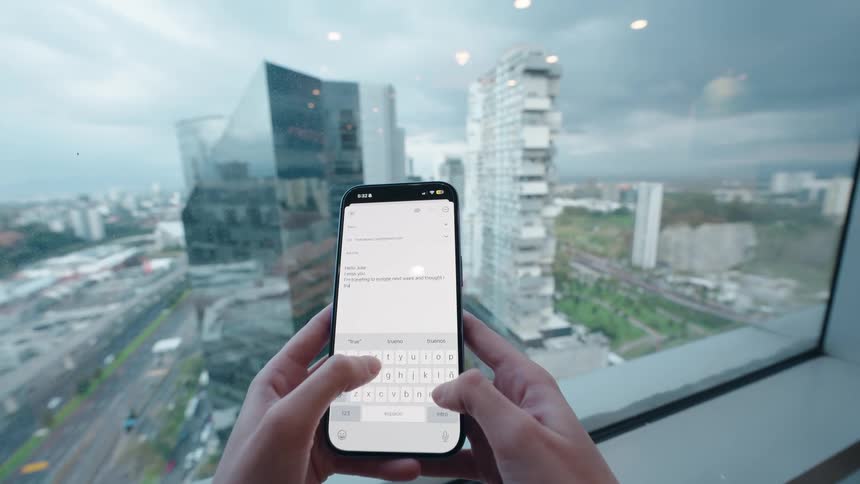 A POV shot of hands typing an email on a smartphone in a high-rise building with a view of a modern cityscape
