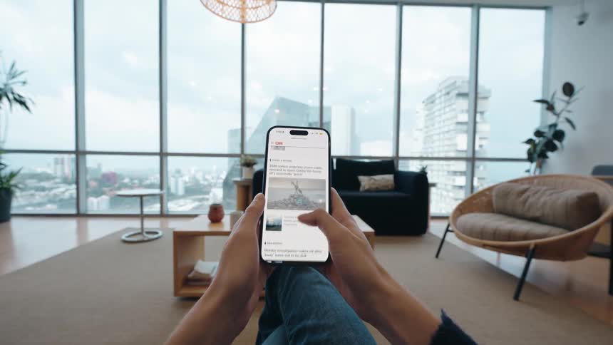 A first-person point of view of a person scrolling through a news feed on their smartphone while relaxing in a modern apartment overlooking the city