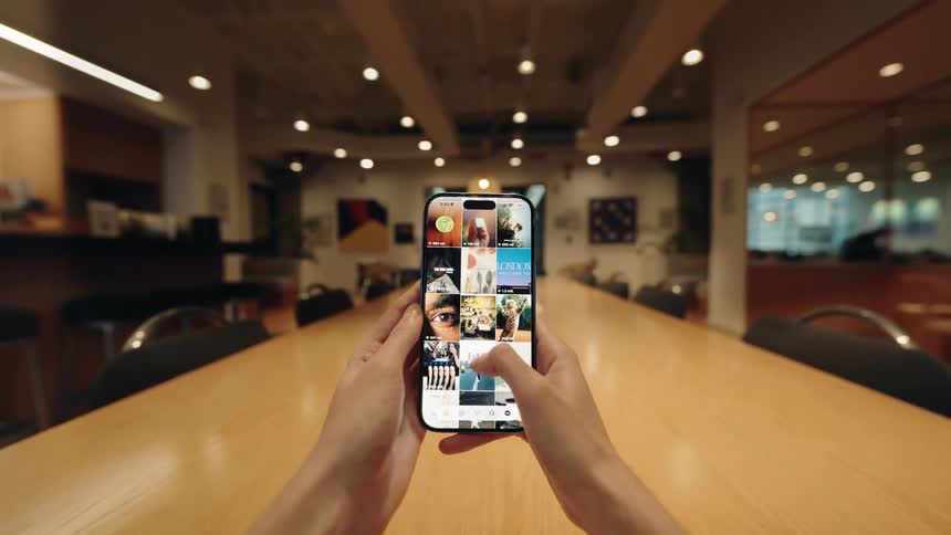 A first-person perspective of hands holding a smartphone and endlessly scrolling through a social media photo feed at a conference table