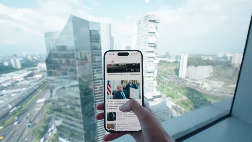 A person scrolls through a news website on their smartphone in a modern high-rise office overlooking the city