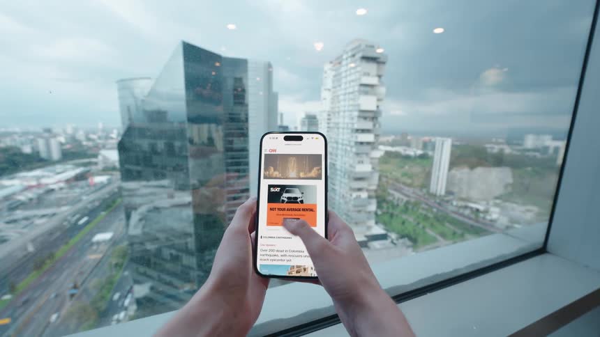 First-person view of hands holding a smartphone and browsing a news feed with a modern cityscape in the background