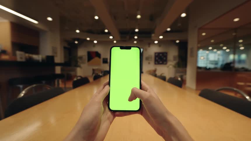 POV of a person using a smartphone with a green screen, swiping and tapping while sitting at a long wooden table in a modern office