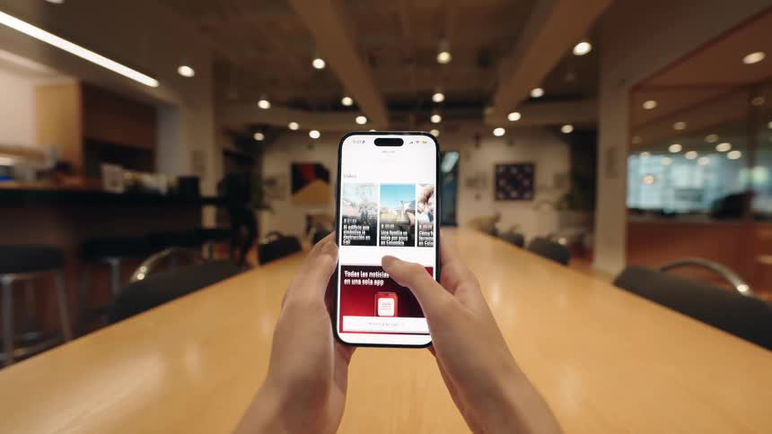A point-of-view shot of a person scrolling through articles and news on a smartphone while sitting at a long wooden table in a modern office