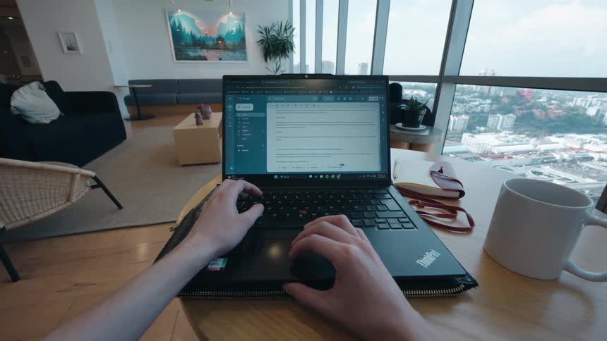 A first-person perspective of a person working remotely on their laptop, typing an email in a modern living room with a panoramic city view