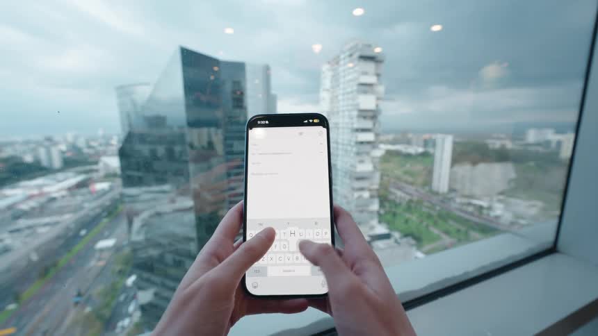 A person holds a smartphone and types an email in a modern high-rise office with a panoramic view of the city