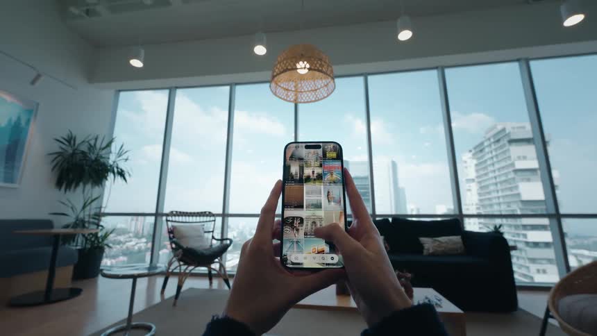 A point-of-view shot of a person scrolling through a social media photo feed on a smartphone in a modern apartment overlooking the city
