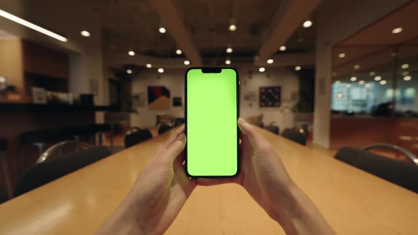 First-person perspective of hands holding a smartphone with a green screen in a modern office conference room