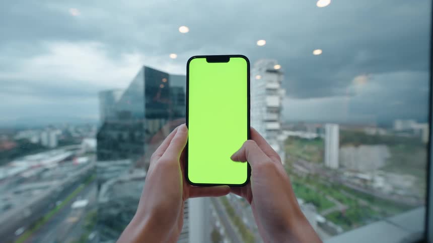 A first-person view of hands holding and swiping on a green screen smartphone in front of a window with a modern cityscape view