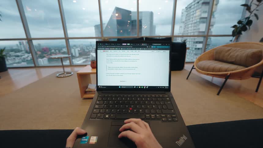 A first-person point-of-view of someone scrolling through a news website on a laptop in a modern high-rise apartment overlooking a city