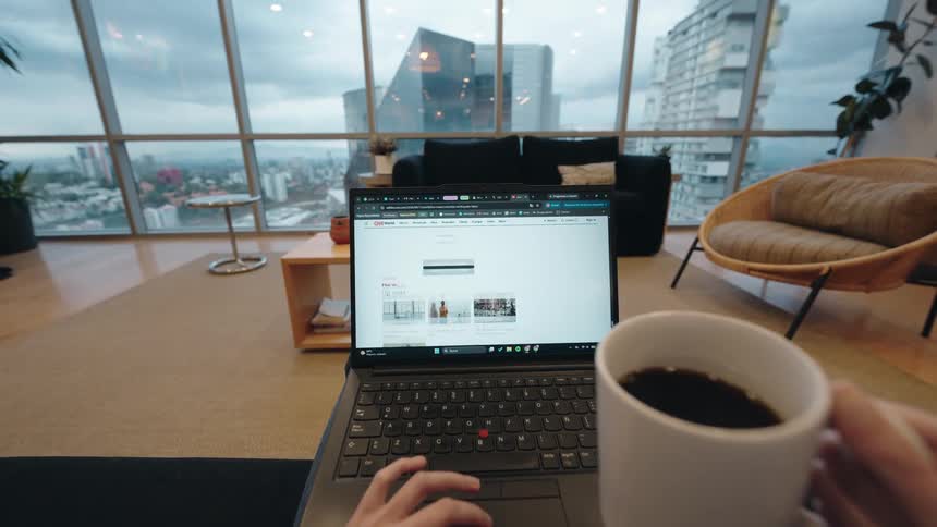 POV of a person enjoying a cup of coffee while browsing news and coding on their laptop in a modern apartment overlooking the cityscape
