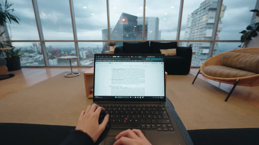 POV timelapse of hands typing on a laptop in a modern penthouse apartment with a city view on a cloudy day