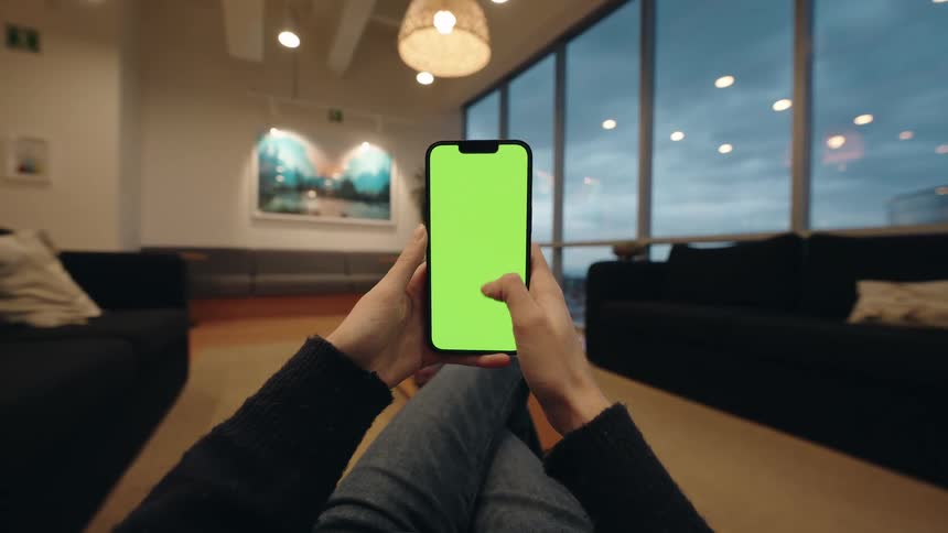 A first-person view of a person relaxing in a modern lounge while scrolling through a smartphone with a green screen