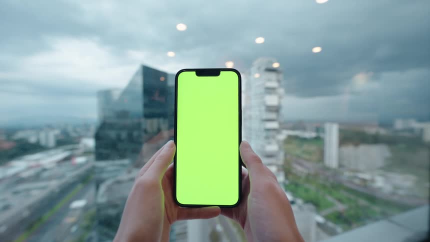 Point of view shot of hands holding a vertical smartphone with a green screen in front of a panoramic window showing a modern city