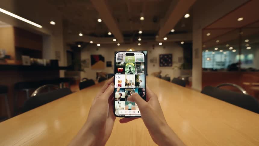 First-person perspective of hands holding a smartphone and scrolling through a social media feed in a modern office