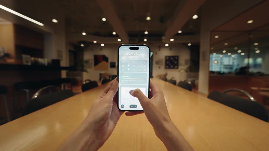 A first-person point of view of a person's hands holding a smartphone and scrolling through a weather forecast app in a modern office space