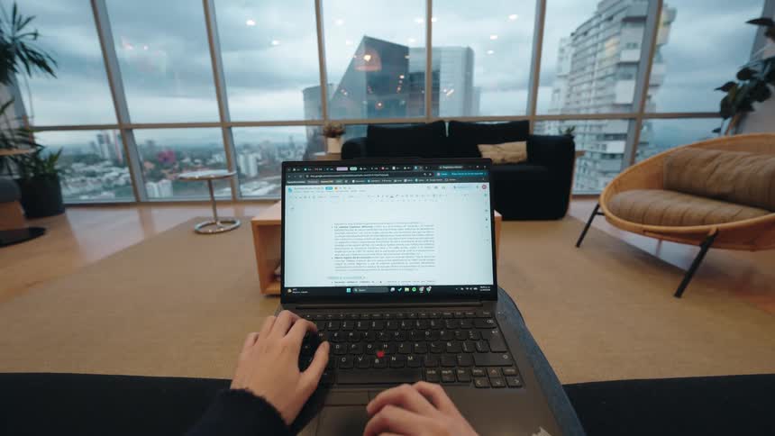 First-person perspective of a person typing on a laptop in a modern high-rise apartment with a large window overlooking the city