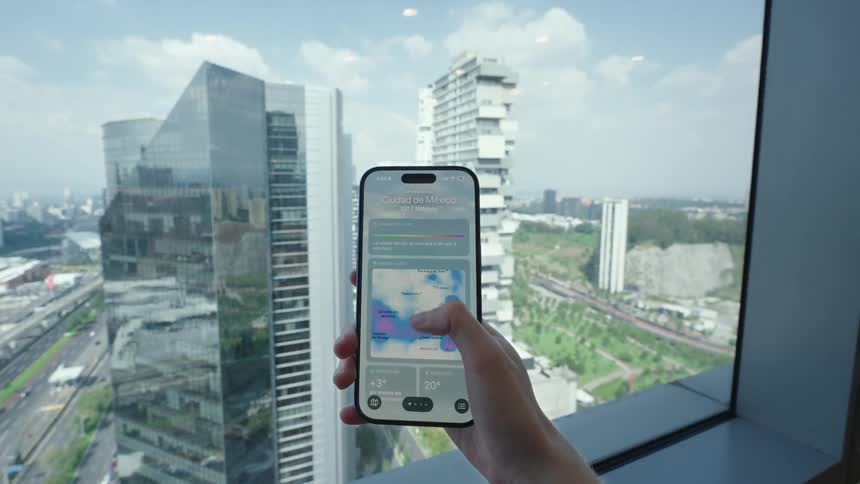 A person checks the detailed weather forecast on their smartphone in front of a window with a sprawling modern cityscape view