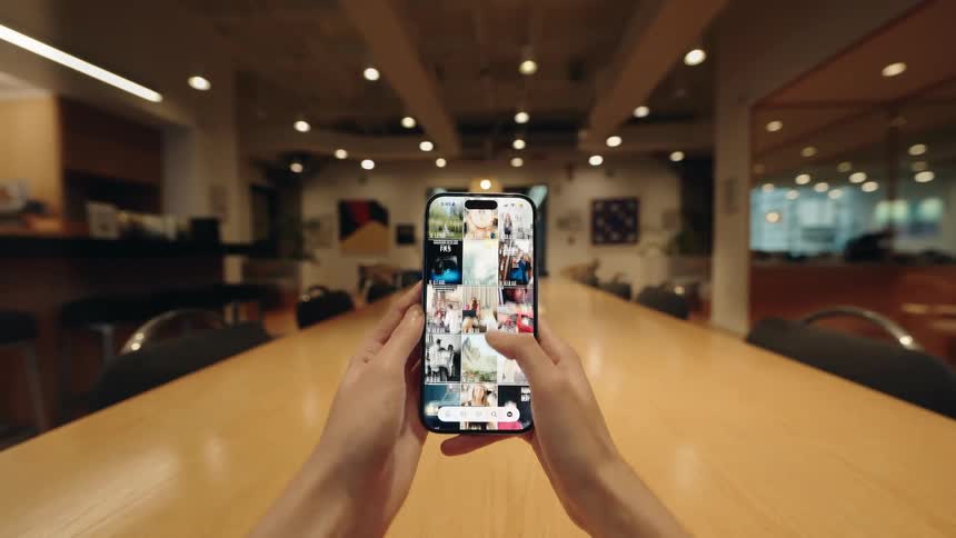 A person's hands scroll through a vibrant social media photo gallery on a smartphone while sitting at a long wooden table in a modern office