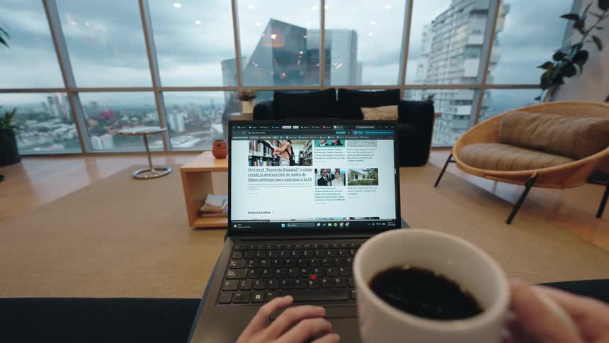 First-person perspective of a person working on a laptop and drinking coffee in a modern apartment overlooking a cityscape