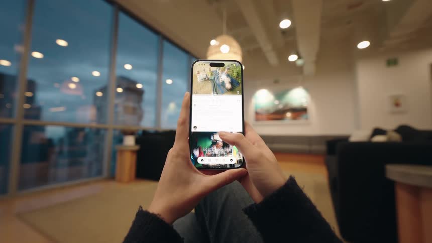 A first-person perspective of hands holding a smartphone and endlessly scrolling through a social media feed in a modern lounge with city views at twilight