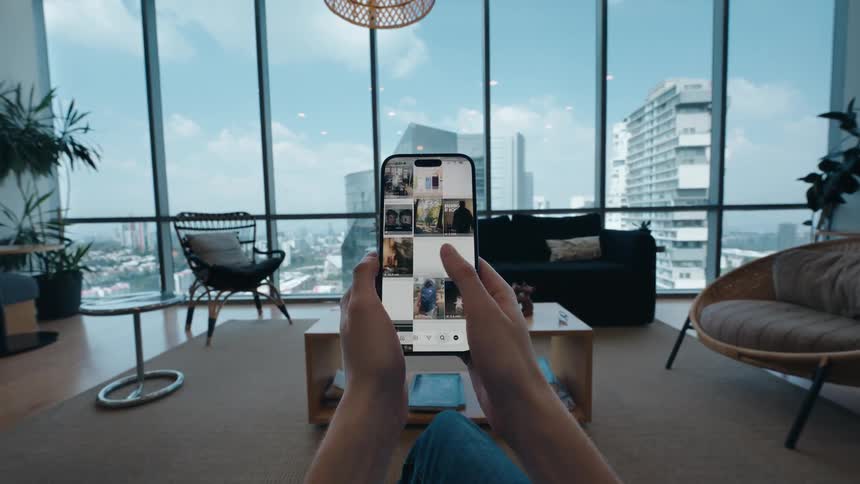 First-person view of someone scrolling through photos on a social media app in a stylish high-rise apartment overlooking the city