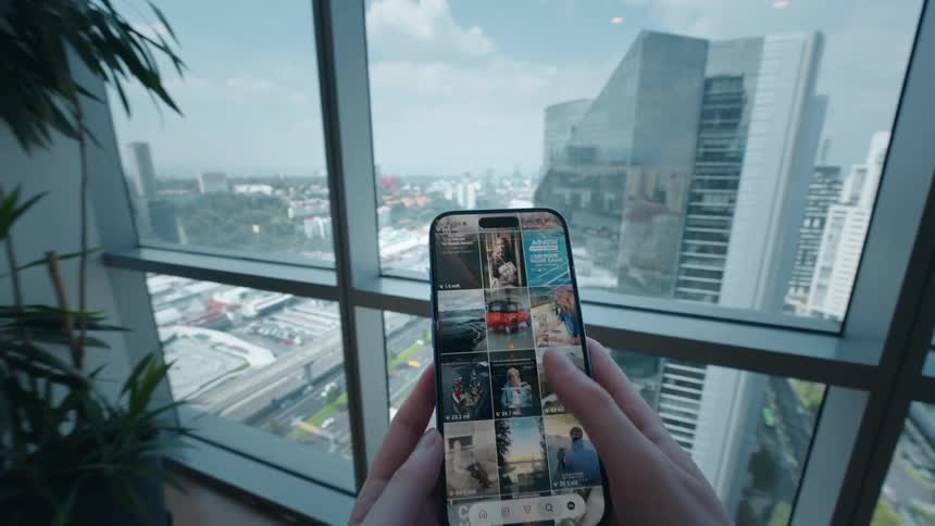 A first-person view of hands scrolling through a social media feed on a smartphone in a modern office with a large window overlooking the city