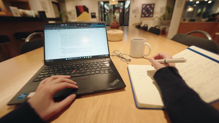 A first-person view of a person working on a laptop and taking handwritten notes in a notebook at a large wooden desk