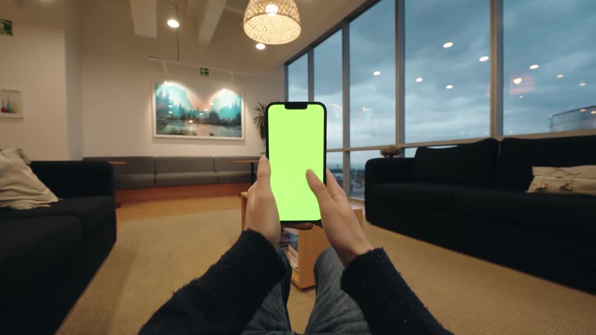 First-person perspective of hands holding and scrolling through a green-screen smartphone in a modern, well-lit lounge area
