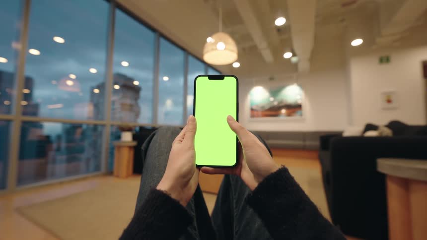 A first-person view of a person relaxing in a modern office lounge while scrolling on a smartphone with a green screen