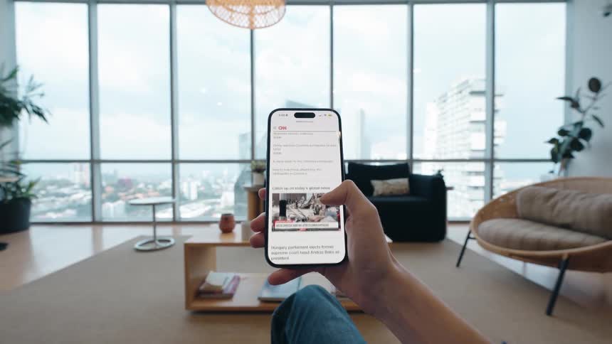 A person scrolls through the CNN news app on a smartphone in a modern apartment with a city view